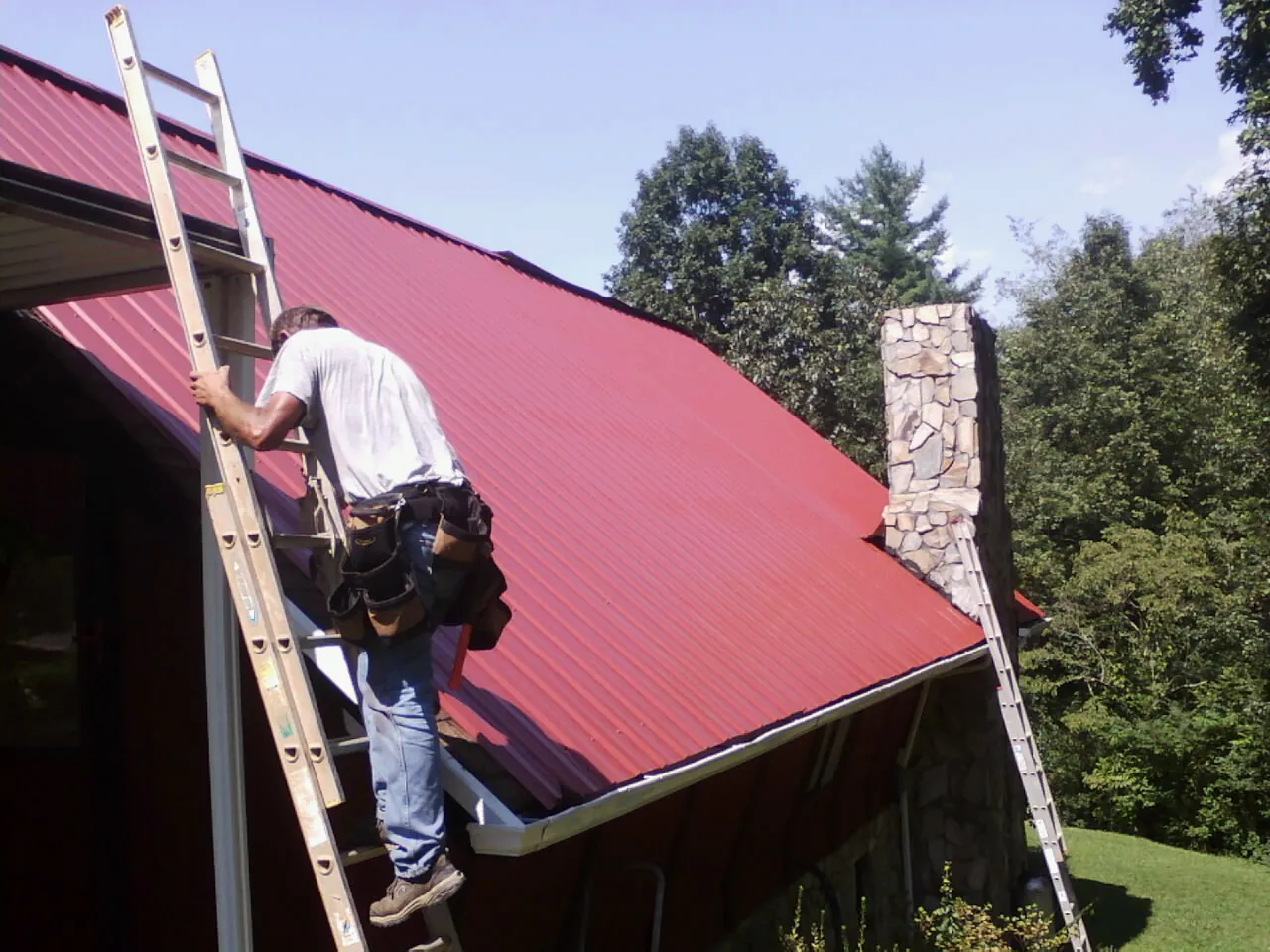 Roofer on a ladder beside a red metal roof and stone chimney — Soffit & Fascia Repair by Roofing Brothers in Gratz